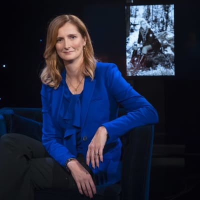 Annica Bresky sitter i tevestudion i kungsblå kavaj och topp med ett svartvitt foto i bakgrunden som föreställer henne  sittandes på en sten i skogen.