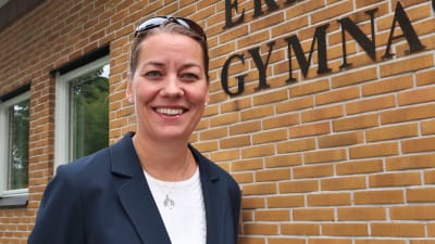 Porträtt av rektor Petra Blomwvist som ser rakt in i kameran och ler. I bakgrunden synns tegelväggen av Ekenäs gymnasiums byggnad och texten "Ekenäs gymnasium" .