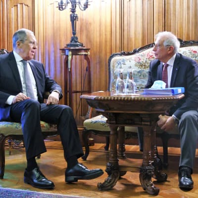 Sergei Lavrov istuu tuolilla ja Josep Borrell sohvalla. Lavrov puhuu juuri, Borrell kuuntelee. Miehillä on tummat puvut, kravatit. Heidän edessään on pieni pöytä, jossa kaksi vesipulloa ja paketti, mahdollisesi kasvomaskeja.