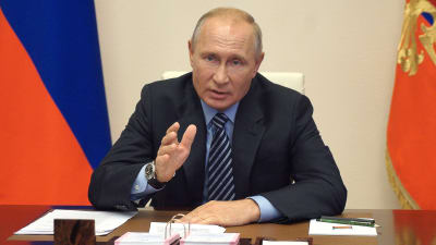 Rysslands president Vladimir Putin sitter vid ett sort skrivbord och pekar med hela handen. Den ryska federationens flagga står i bakgrunden.