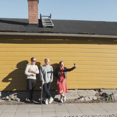Kolme henkilöä nojaa keltaiseen lautaseinään ja ottaa selfie-kuvaa yhdessä (kuvassa Henri Ylikulju ja Egenland-ohjelman juontajat Nicke Aldén ja Hannamari Hoikkala)