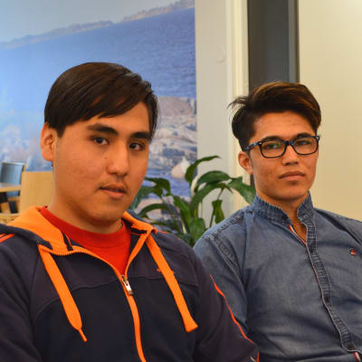 17-åringarna Ezzat Najafi och Masaud Alizada sitter på en soffa i deras boende, i bakgrunden korridor, krukväxter och matbord.