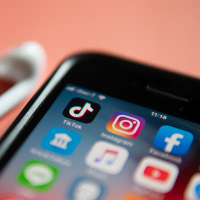 En telefon och ett par hörlurar ligger på ett persikofärgat bord. På telefonen syns olika appar, bland annat Tiktok och Instagram.