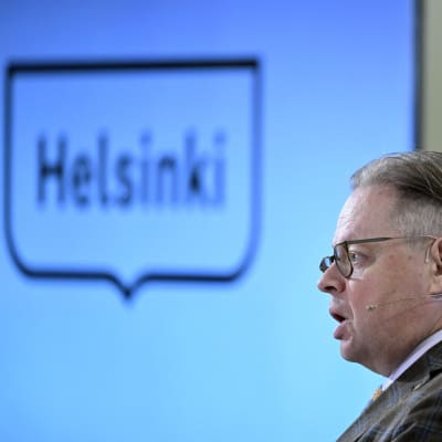 Borgmästare Juhana Vartiainen står framför en skärm med Helsingfors stads logga.