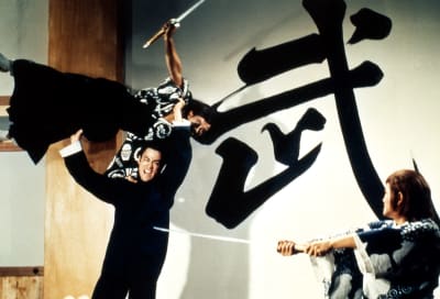 Mustiin pukeutunut Bruce Lee on nostanut kimonoasuisen miekkamiehen ilmaan ja on paiskaamassa häntä kohti toista japanilaissoturia. Kuva elokuvasta Vihan nyrkki.