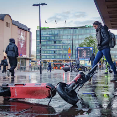 Kaatuneena maahan jätetty sähköpotkulauta märällä kiveyksellä. Taustalla ihmisiä.