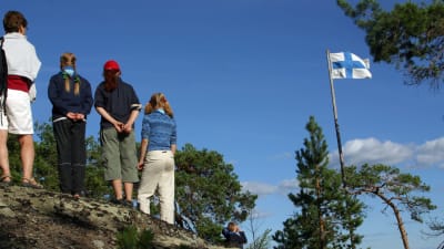 Nuoria partiolaisia seisoo korkealla kalliolla. Oikealla puolella Suomen lippu salossa. 