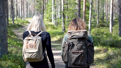 Fotade bakifrån går Marina och Lina i en grön skog med ryggväskor.