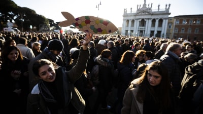 En stor folksamling på ett torg i Rom, en person i förgrunden med en sardin gjord av papp i handen. 