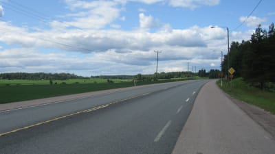 En asfalterade väg med åker på ena sidan och träd på andra. Trafikmärke för förkörsrätt. Telefonstolpar. Sommar, blå himmel med moln.