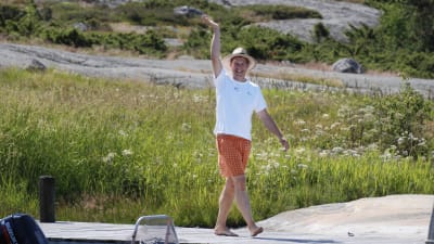Riko Eklundh går på en brygga och vinkar. Han har orangerutiga shorts, vit t-skjorta och en halmhatt på sig.