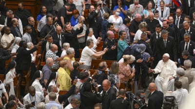 Påven i rullstol rullas upp för mittgången i en katedral och hälsar på människor i närheten. Många i kyrkbänkarna sträcker fram mobiltelefoner och TV-kameror finns också på plats.