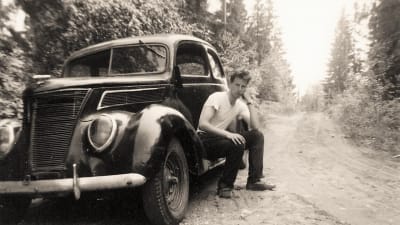 Svartvit bild av en ung man som sitter vid sin bil på en skogsväg.
