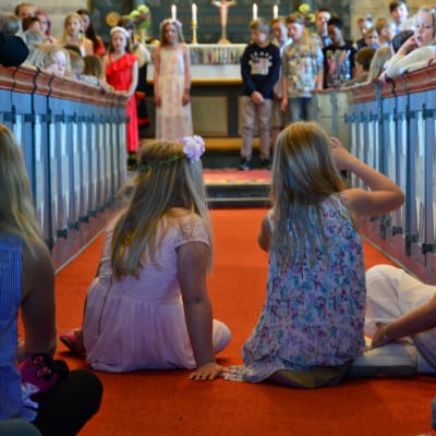 Några flickor sitter i altargången i en kyrka. Uppe vid altaret står uppklädda skolelever.