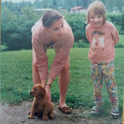 Äiti ja tytär ovat vaaleanpunaisissa vaatteissa nurmikolla pienen koiranpennun kanssa.