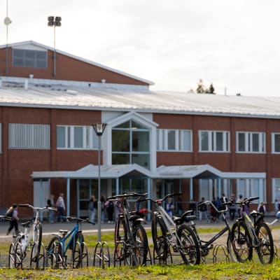 Koulun piha loppukesällä. Taustalle punaruskeatiilinen koulurakennus ja sen edessä koululaisia. Edustalla polkupyöriä telineessä.