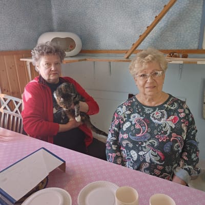 Två kvinnor sitter vid ett kaffebord. Ena kvinnan håller i en katt. 