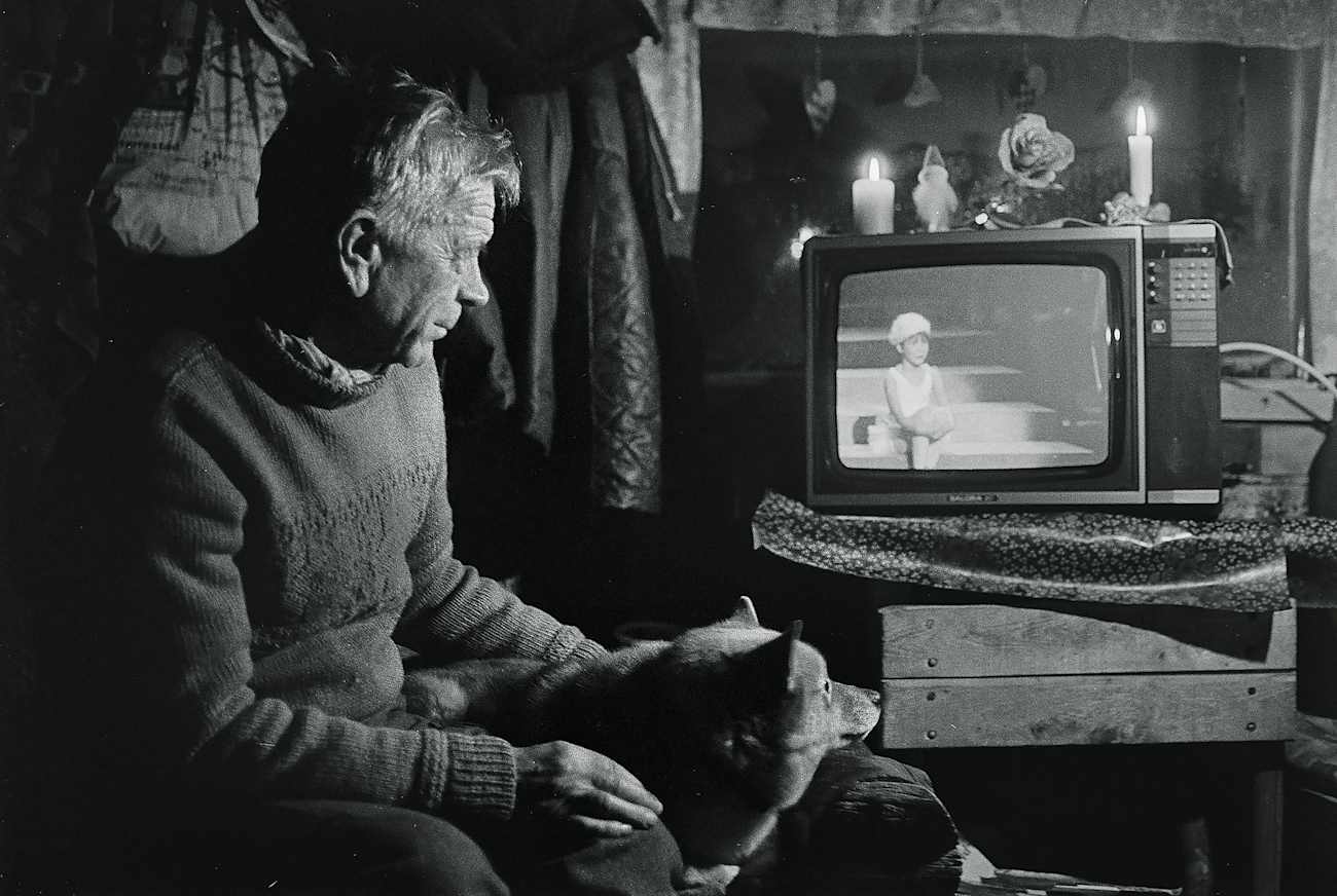 Mies katsoo televisiosta ohjelmaa, vierell��n koira, kodikkaassa ja h�m�r�sti valaistussa huoneessa.