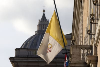 Vatikanens flagga vajar på halv stång utanför ett hus med Peterskyrkans kupol i bakgrunden.