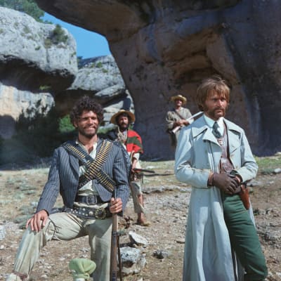 Kivääriä ja asevyötä kantava mies sekä vaaleampi pitkään takkiin pukeutuva mies katsovat samaan suuntaan, kaksi asemiestä ja kalliomaisema takanaan.