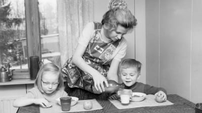 En mamma häller upp mjölk åt sina barn vid matbordet.