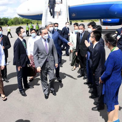 Mies, Kiinan ulkoministeri Wang Yi, harmaassa puvussa astuu ulos lentokoneesta.  Vastassa rivi burmalaisia henkilöitä.