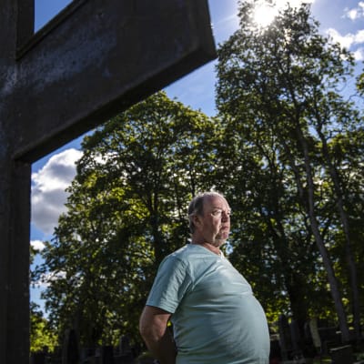En man står med händerna bakom ryggen framför ett stort kors på en kyrkogård.