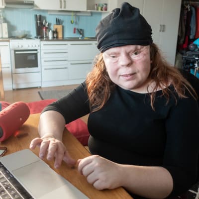 En kvinna sitter hemma framför en dator. Till höger, från kvinnan sätt, finns en orange högtalare. 