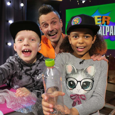 Superhjälparnas programledare Jontti Granbacka och två barn ler glatt, flickan håller upp en plastflaska mot tittaren