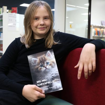 10-vuotias Armi Yoki istuu kirjaston sohvalla pitäen kädessä kirjoittamansa Miu Miu Karvapallo -kirjaa, jonka kannessa on pörröinen kissanpentu.