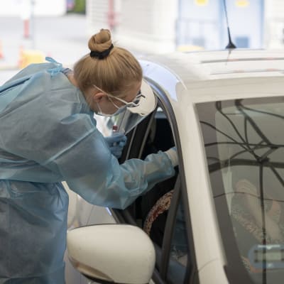 Närvårdare i skyddsdräkt tar coronaprov av klient sittande i bil. 