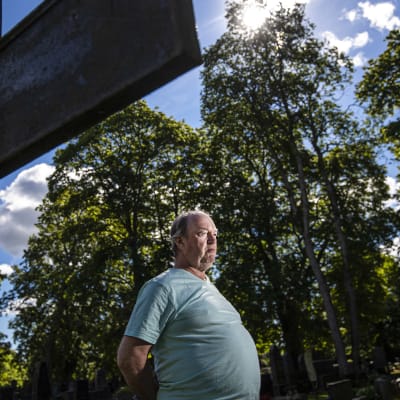 En man står med händerna bakom ryggen framför ett stort kors på en kyrkogård.
