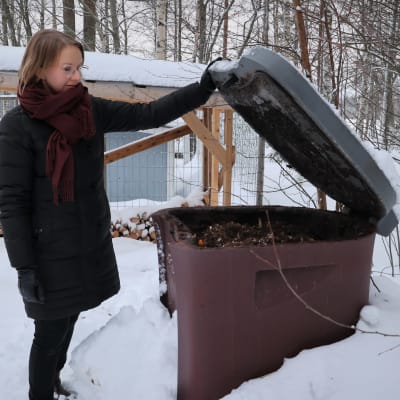 Nainen nostaa kompostin kantaa lumisella pihalla.