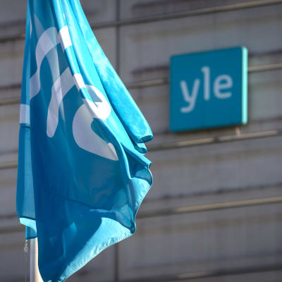 Yleisradio-logotyp på en flagga och på en gråhusvägg i bakgrunden. 