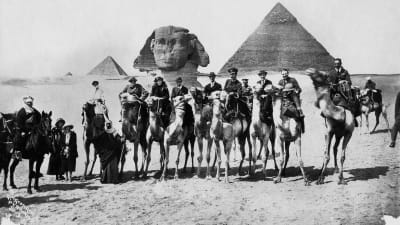Svartvid bild av sällskap på kameler vid pyramiderna. På bilden syns bland nadra Gertrude Bell flankerad av Winston Churchill och T.E. Lawrence.