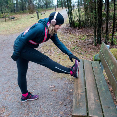 Liisa-Maija Huttunen i träningskläder och reflexsele. Hon står och stretchar benet mot en bänk i en skog.