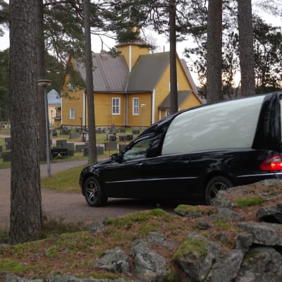 En svart begravningsbil kör in på kyrkogården i Dragsfjärd
