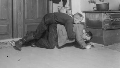 Två män leker "ryggbördan", där den ena ligger på golvet med den andra på sin rygg, Den undre ska försöka stiga upp, den andra ska hindra den första att göra det.