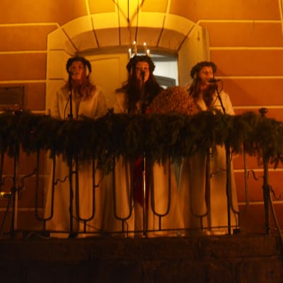Lucia och två tärnor står utomhus på en trappa och sjunger i mikrofoner.