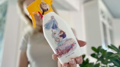 En kvinna står och håller fram en flaska med solkräm som skymmer hennes ansikte. På flaskan finns fastlimmade klistermärken på Frozen-karaktärer.