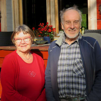 En äldre kvinna och äldre man står framför en rödmålad stockstuga. De ser in i kameran och ler.