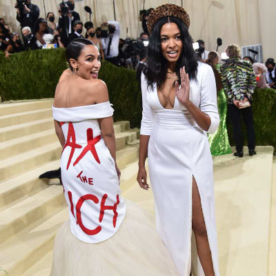 Alexandria Ocasio-Cortez på Metgalan iklädd en vit klänning där det står "Tax the rich" med röda bokstäver, bredvid henne Aurora James som designat klänningen.