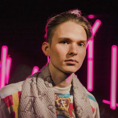 Vaaleahiuksinen nuori mies katsoo suoraan kameraan vakavalla ilmeellä pinkkien neonvalotolppien edessä.