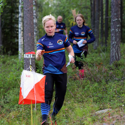 Vaaran perhe juoksee suunnnistusvarusteissa lehtimetsässä, kuvassa näkyy myös rastin merkki.