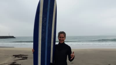 Malin Kivelä står med surfbräda och en vid ocean i grådaskigt ljus i bakgrunden. Bär våtdräkt och ler. Tummen uppe.