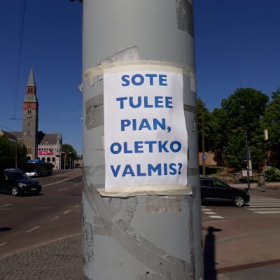 Ett papper med texten "Sote tulee, oletko valmis?" fasttejpad på en lyktstolpe i Helsingfors.