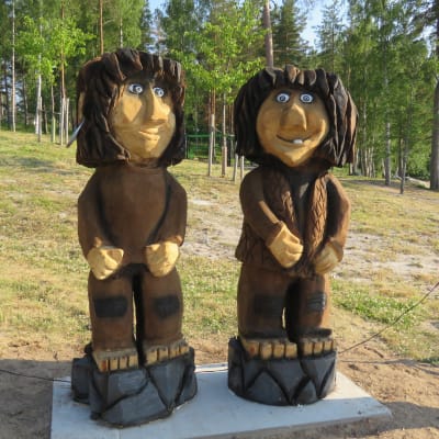 Trästaty  som föreställer två troll gjorda av trä står vid en grässluttning invid vatten och en strand i Bromarv skärgårdshamn.