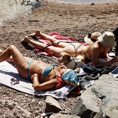 Två kvinnor ligger på handdukar på en strand och solar i bikinis.