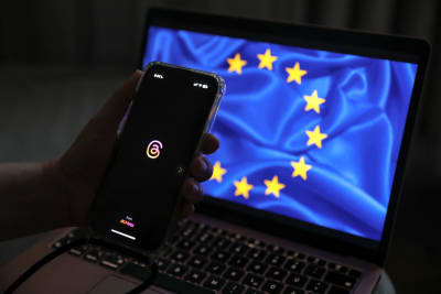 Smartelefon med Threads logga hålls upp framför en dator med EU:s flagga