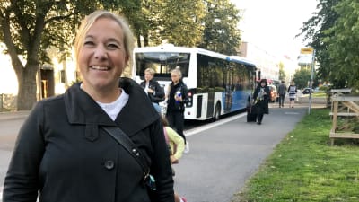 Silja Lindblad står vid busshållplats, i bakgrunden människor och buss.